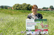 Frau steht in Blühfläche und hält Schild mit Aufschrift "Lebensräume verbessern"